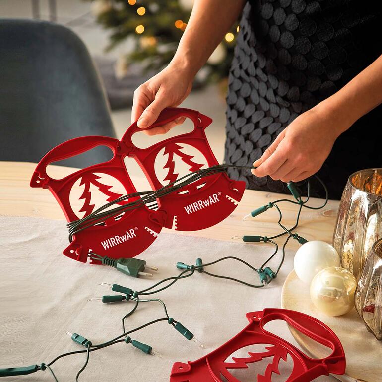 Dankzij het gepatenteerde insteeksysteem kunnen de opbergers afzonderlijk of samen worden gebruikt. Persoon wikkelt kerstverlichting op met WIRRwAR-oproller op een tafel.