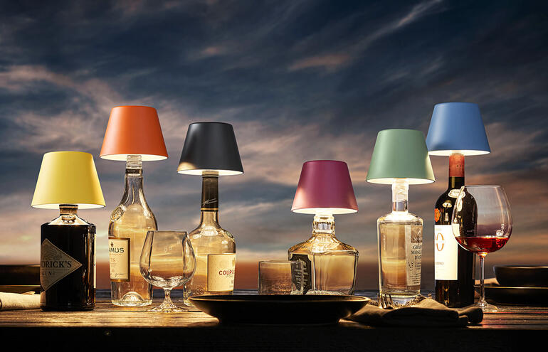 De led-lampenkappen veranderen al uw decoratieve flessen (leeg of nog vol) in bijzondere tafellampen. Zes LED-accu-fleslampen in verschillende kleuren op een tafel.