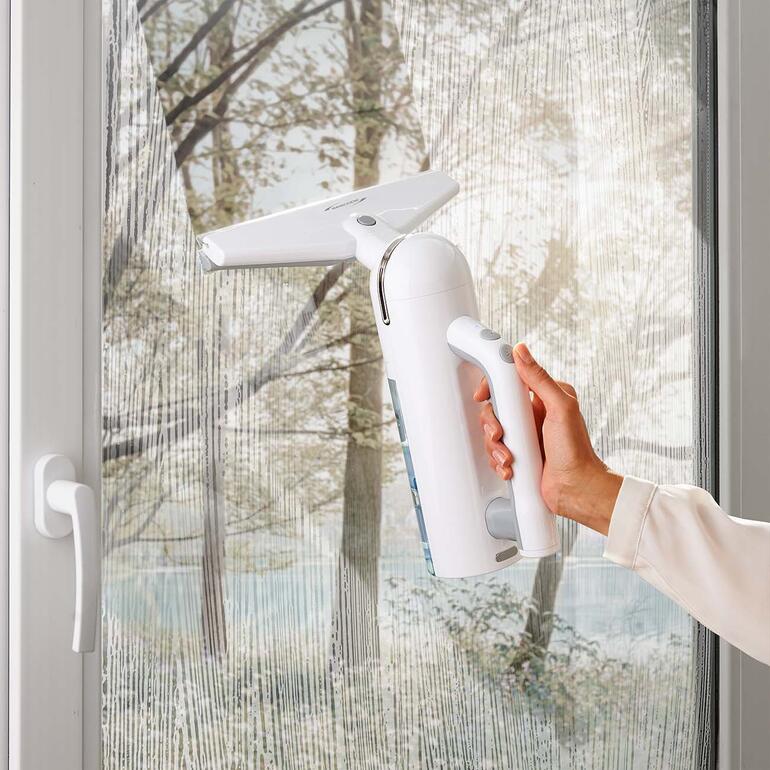 Hand houdt een witte accu-vensterreiniger voor een raam.