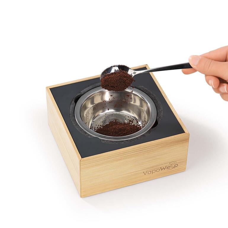 Gewoon in de edelstalen vuurschaal 1 theelepel kruidenmengsel vermengen met 2 theelepels koffiepoeder, deze mix vervolgens aansteken en de box sluiten. VapoWesp rookdoos met een hand die koffiepoeder toevoegt.