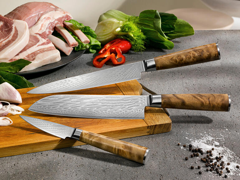 Drie damastmessen op een snijplank met ingrediënten op de achtergrond