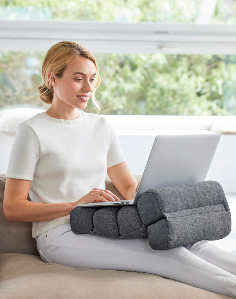 Ideaal als comfortabel rugkussen en als steun voor uw wervelkolom tijdens het werken, autorijden,... Vrouw zit met flexi-rolkussen dat haar armen steunt, achter de laptop op de bank.