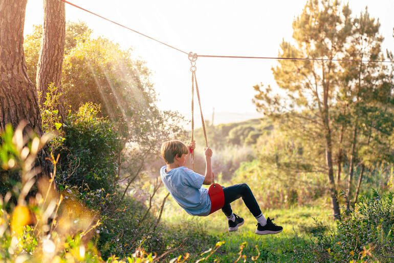 Zweven van boom naar boom aan de zipline op het in hoogte verstelbare schommelzitje is ontzettend leuk. Jongen hangt op een touw in een outdoorparcours bij zonsondergang.