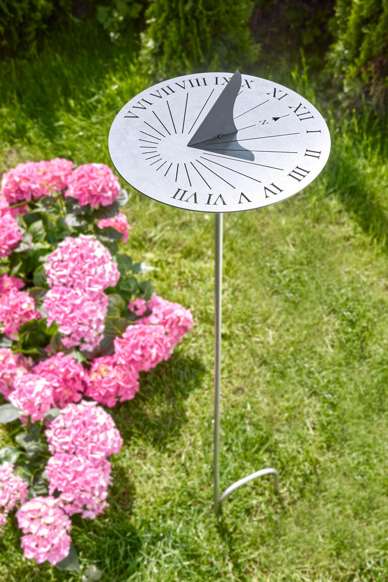 RVS zonnewijzer op een groen gazon, omgeven door roze bloemen.