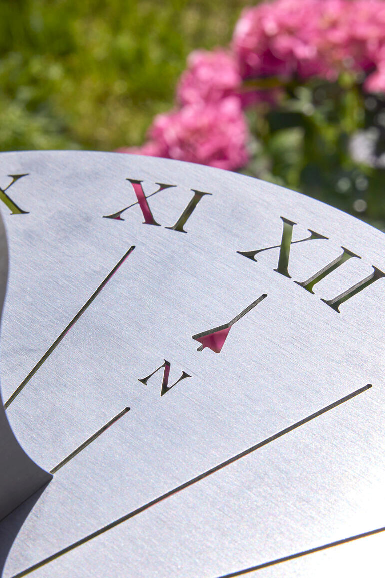 De ring met stralen en cijfers zijn nauwkeurig met een laser in de matte edelstalen schijf gebrand. Close-up van de cijfers op een RVS zonnewijzer met roze bloemen op de achtergrond.