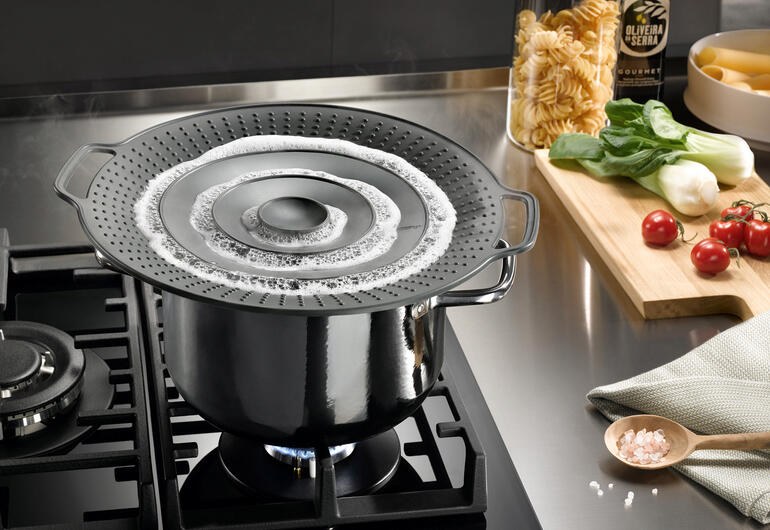 Pan met spatdeksel op hetfornuis, pasta en groenten ernaast.