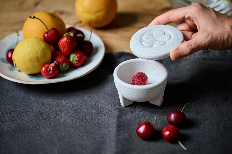 Porseleinen fruitvliegenval met een hand die het deksel opent en fruit op tafel.