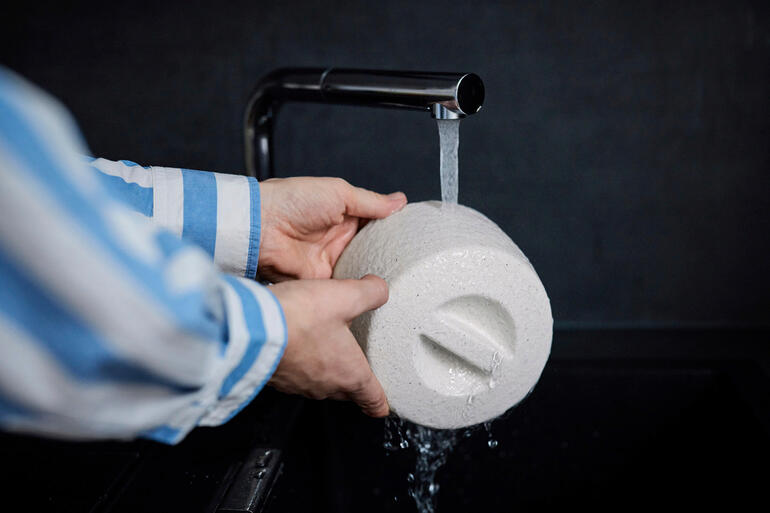 Week de keramische boterkoeler eenvoudig onder stromend water. Het verkoelende effect ontstaat door de verdamping van het geabsorbeerde water. Persoon wast keramische botervorm onder stromend water.