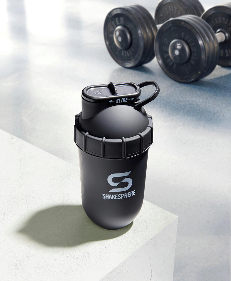 Deze fitness-shaker gebruikt alleen de in de afgeronde capsulevorm ontstane middelpuntvliedende kracht – zonder hoeken waarin poederresten zich kunnen vastplakken. Zwarte shaker op een witte ondergrond met gewichten op de achtergrond.
