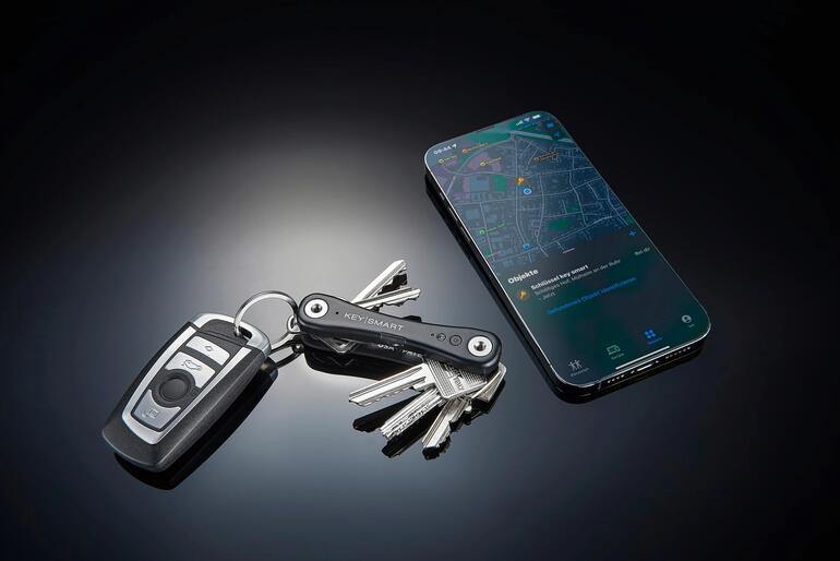 Als uw KeySmart® iPro gelokaliseerd is, ziet u de positie in de app ‘Zoek mijn’ op uw iPhone. Slimme sleutelhanger naast een sleutelhanger en een smartphone op een tafel.