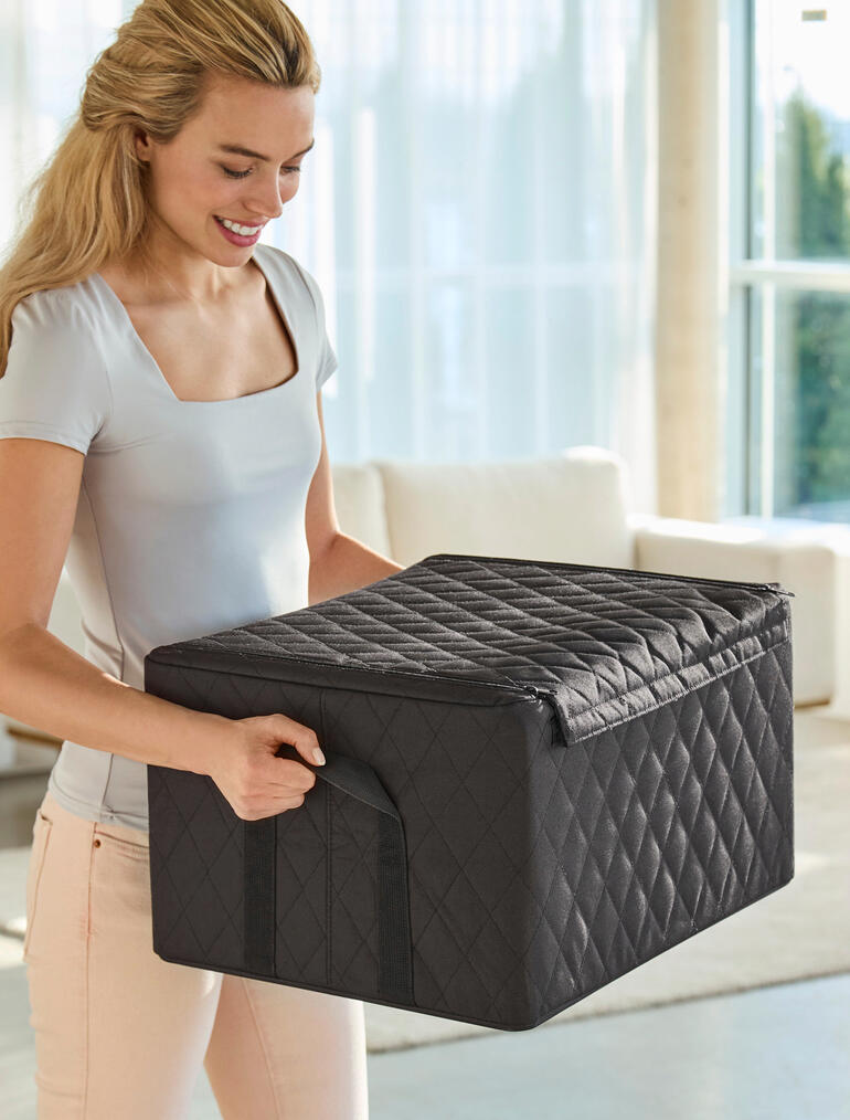 Stevige handgrepen voor comfortabel en veilig vervoer. Vrouw heft een zwarte stapelbox met gestikt ruitpatroon, in de woonkamer.