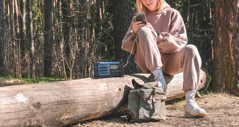 Vrouw in beige kleding zit op een stam met een radio en een rugzak.