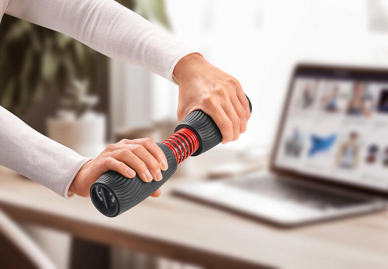 Persoon traint met een pols- en onderarmtrainer aan een tafel met laptop.