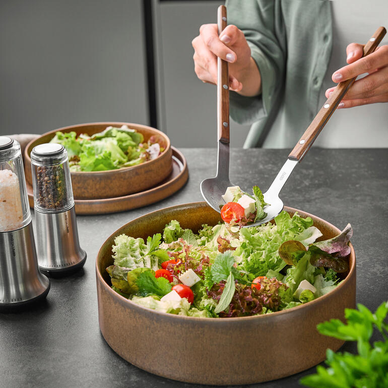 Persoon tilt XL-saladebestek boven een kom met salade.