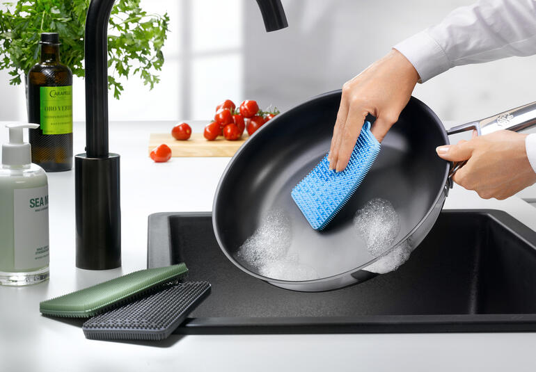 Persoon reinigt een pan met een blauwe siliconen afwasborstel.