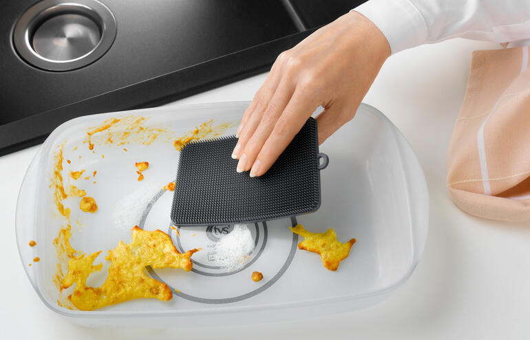 Met de 2 mm lange, harde borstelharen aan de achterkant schrobt u ingebrande etensresten er zo vanaf. Hand gebruikt een zwarte siliconen afwasborstel in een platte schaal.