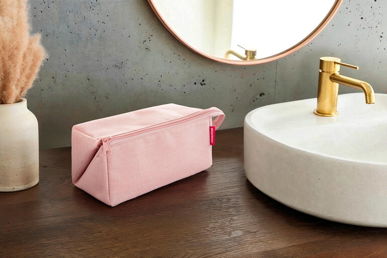 Roze 2-in-1 reisnecessaire op een houten tafel naast een wastafel