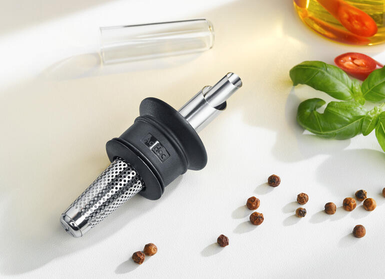 Viervoudig praktisch: de multifunctionele schenktuit doseert, filtert, vangt druppels op en sluit aromatisch af. Decoratieve schenker voor olie en azijn op een witte ondergrond met ingrediënten.
