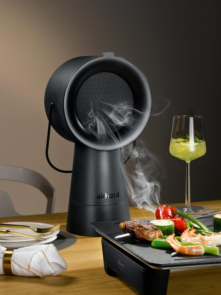 Airhood 2.0 in het zwart, rokend grillapparaat op tafel met groenten en vlees.
