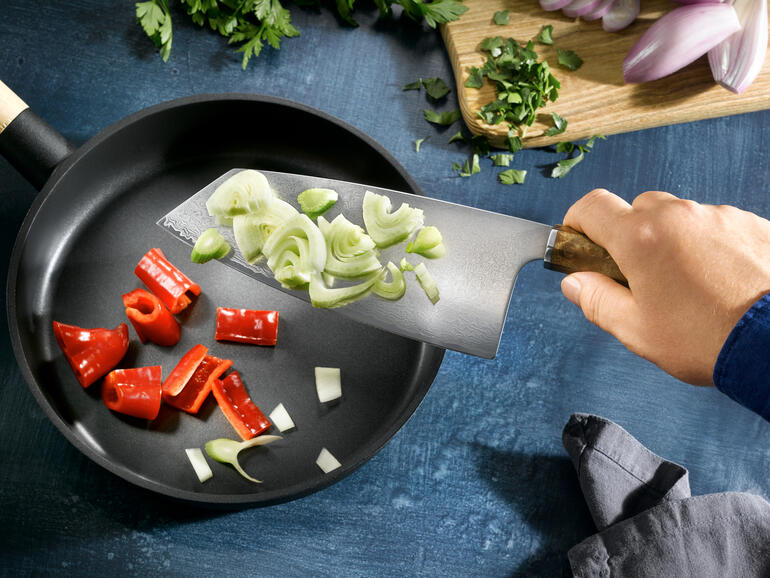 Leg de gesneden etenswaren op het brede lemmet en doe ze zonder knoeien in een pan. Hand met Damast Nakiri-mes die groenten in een pan snijdt.