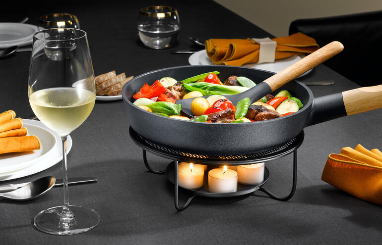Pan met groenten op een vuurplaat, naast een wijnglas op een gedekte tafel.