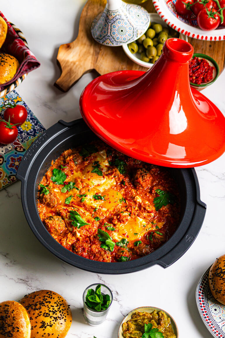 Tajine Peugeot Saveurs met een rode deksel, gevuld met kleurrijke groenten en kruiden, op een tafel met bijgerechten.