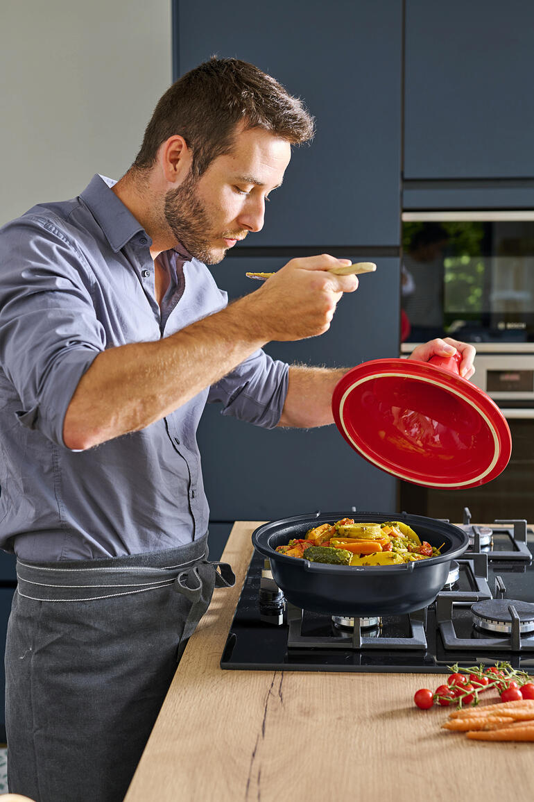 Man in de keuken die de Peugeot Saveurs tagine proeft met een vork terwijl hij de deksel opheft.