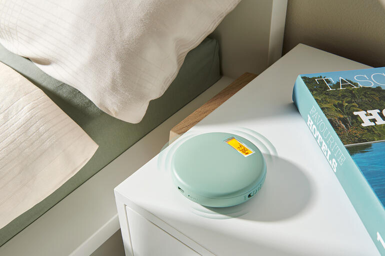 Vibratiewekker in zachtgroen, op een nachtkastje naast een boek en een dekbed.