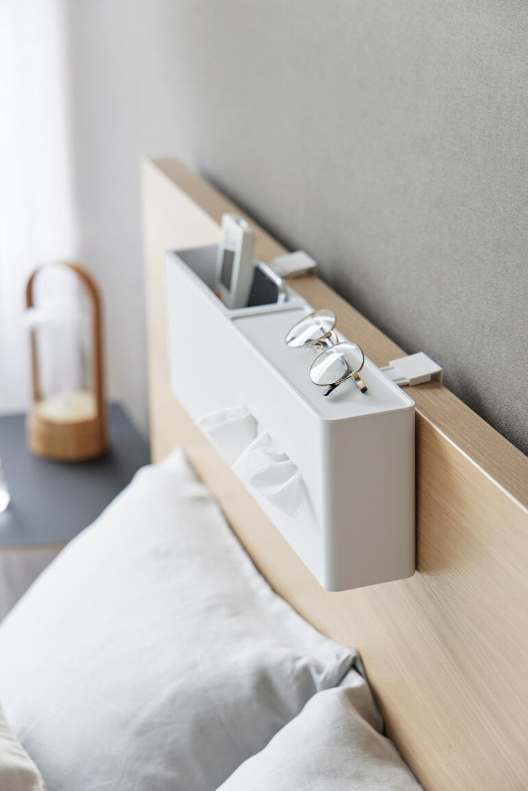 Witte tissuebox aan het hoofdeinde van een bed met bril en smartphone.