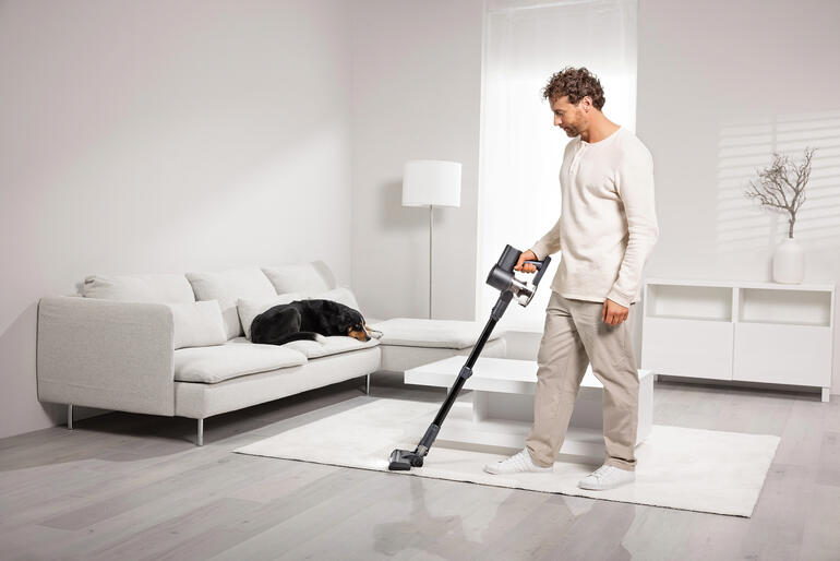 Man zuigt met een draadloze stofzuiger op een tapijt, een hond ligt op een bank.