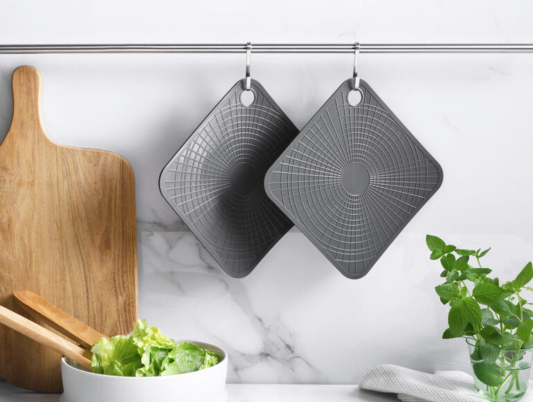 Twee grijze silicone ovenwanten hangen aan een stang in een moderne keuken.