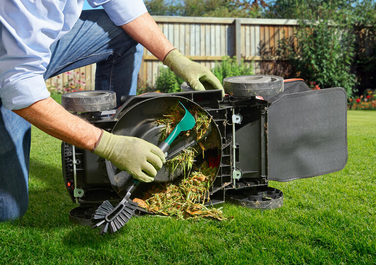 Persoon maakt een omgedraaide grasmaaier schoon met een gereedschap terwijl grasresten worden verwijderd.