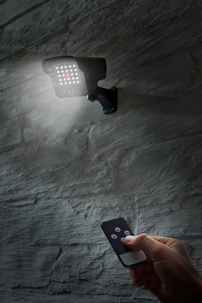 Persoon met afstandsbediening activeert een solar camera imitatie die aan een grijze muur is bevestigd.