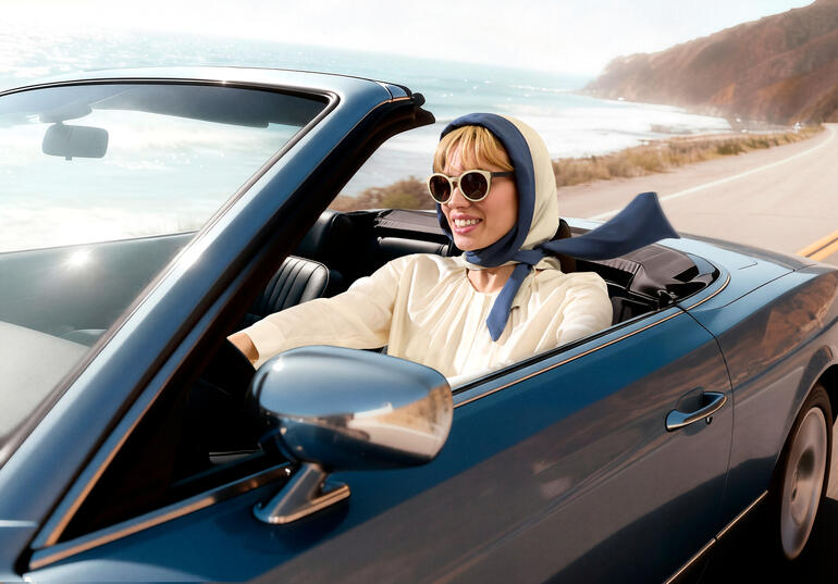Vrouw in een cabrio met hoofddoek en zonnebril rijdt langs de kust.