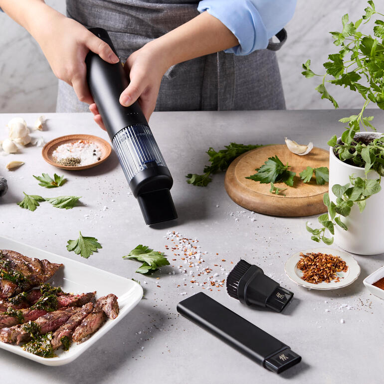 Draadloze XTEND handstofzuiger wordt boven een stuk vlees gehouden, met verse kruiden en specerijen ernaast.