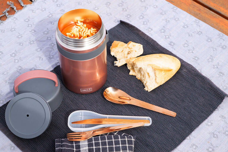 Geïsoleerde snackbox in rosé met geopend deksel, naast brood en bestek op een deken.