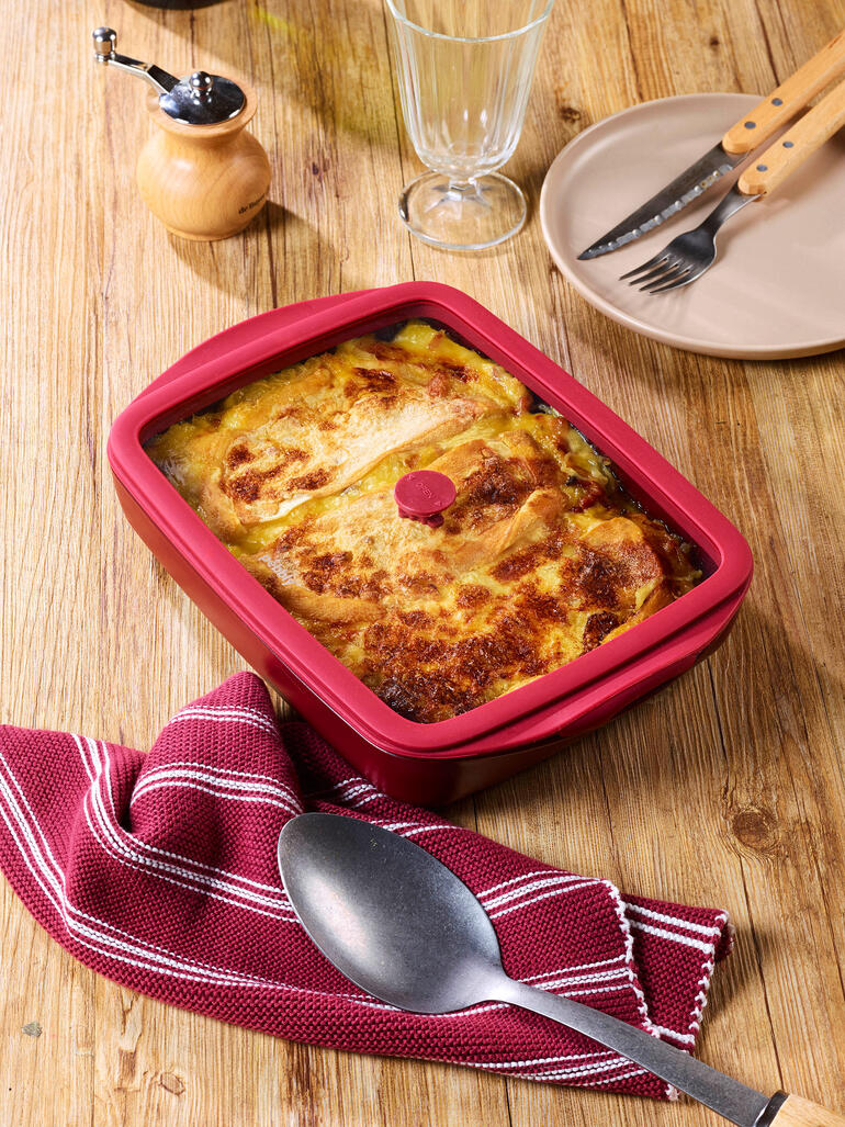 Rode ovenschaal met deksel, gevuld met bonenschotel, op een houten tafel.