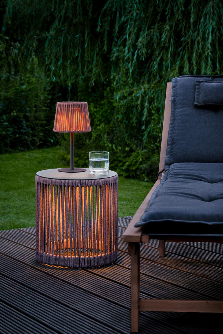 LED-buitentafel met oranje lamp en glas water op houten veranda naast een ligstoel.