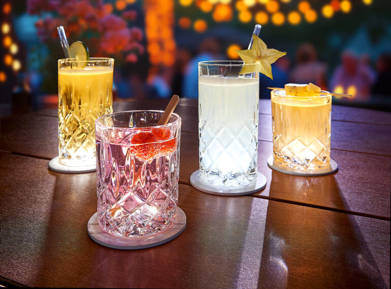 Vier verlichte glazen op een tafel met LED-onderzetters, ideaal voor een feestelijke sfeer.