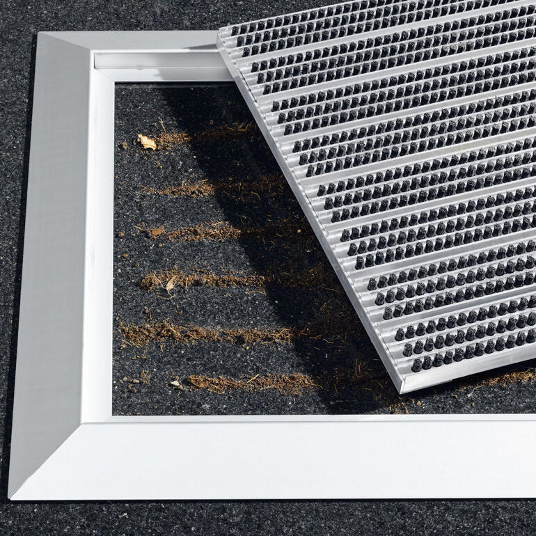 Om de mat schoon te maken tilt u gewoon het borstelrooster op en zuigt of veegt u het vuil weg. Aluminium entree mat met verwijderbare bovenplaat op een vloer.
