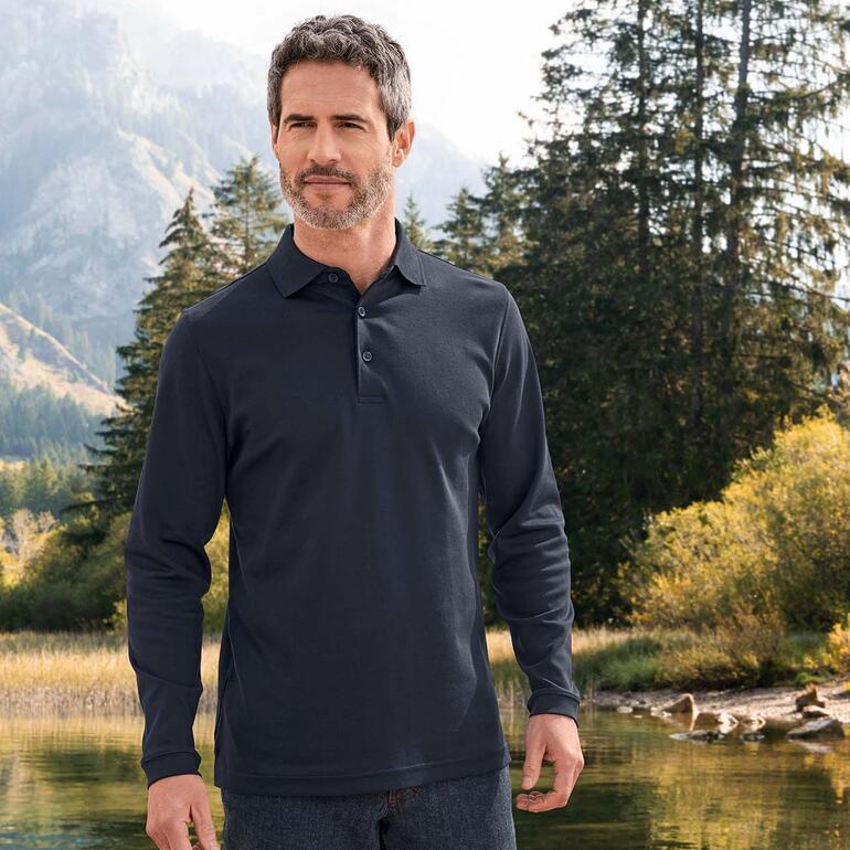 Man in een zwart Pima-interlock-poloshirt, staand in een natuurlijke omgeving met bergen op de achtergrond.