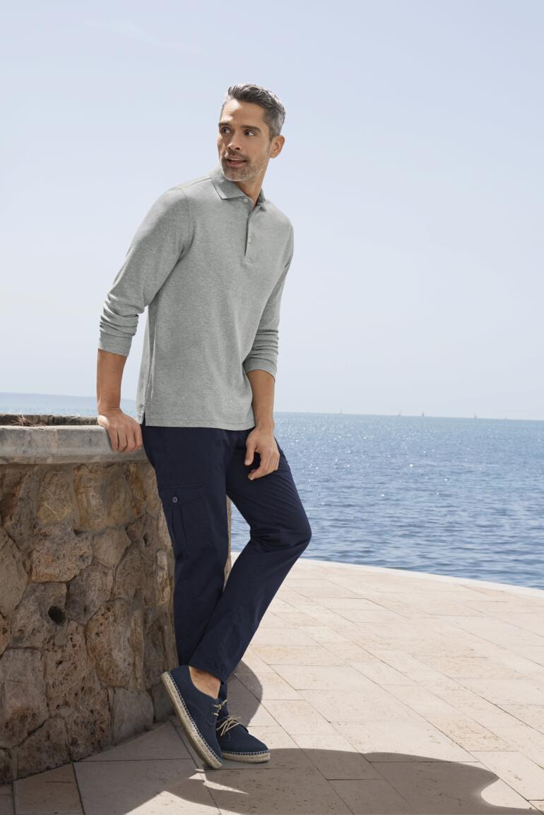Cargo-bermudas Sun Reflect Man in een grijze Pima-interlock polo, staand bij een stenen muur met uitzicht op de zee.