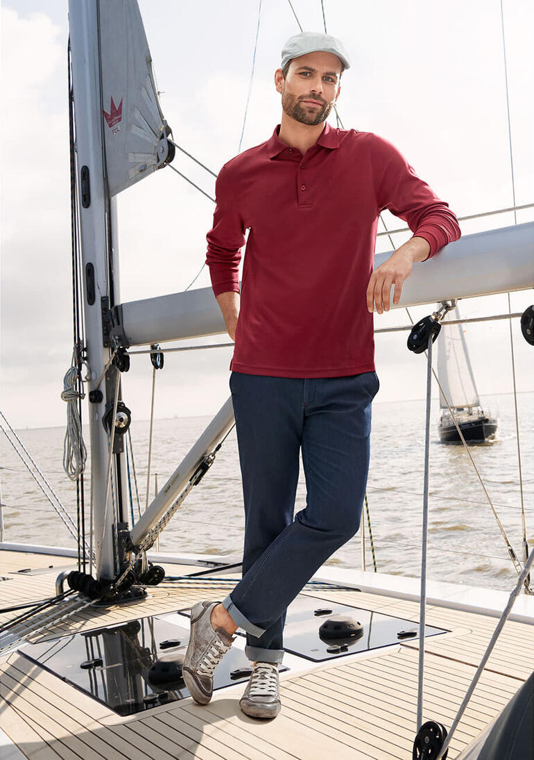 Jeans met zijde Man in een rood Pima interlock polo shirt, staand op een boot, lachend.