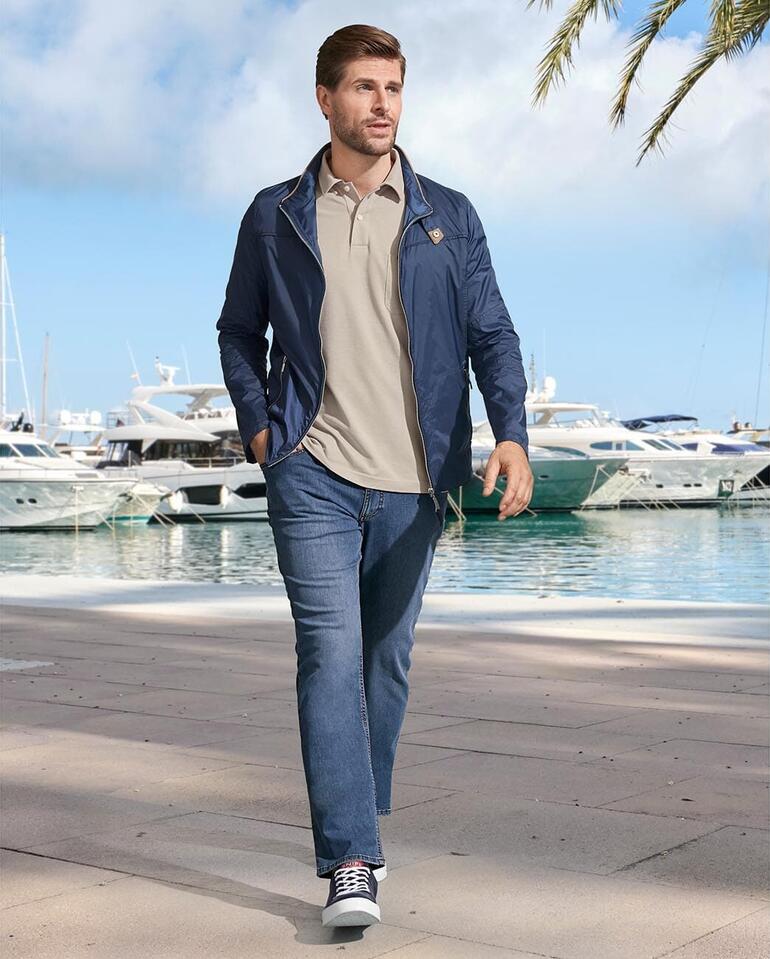 Eurex by Brax zomer jeans Man in een blauwe jas en beige polo loopt langs de haven.