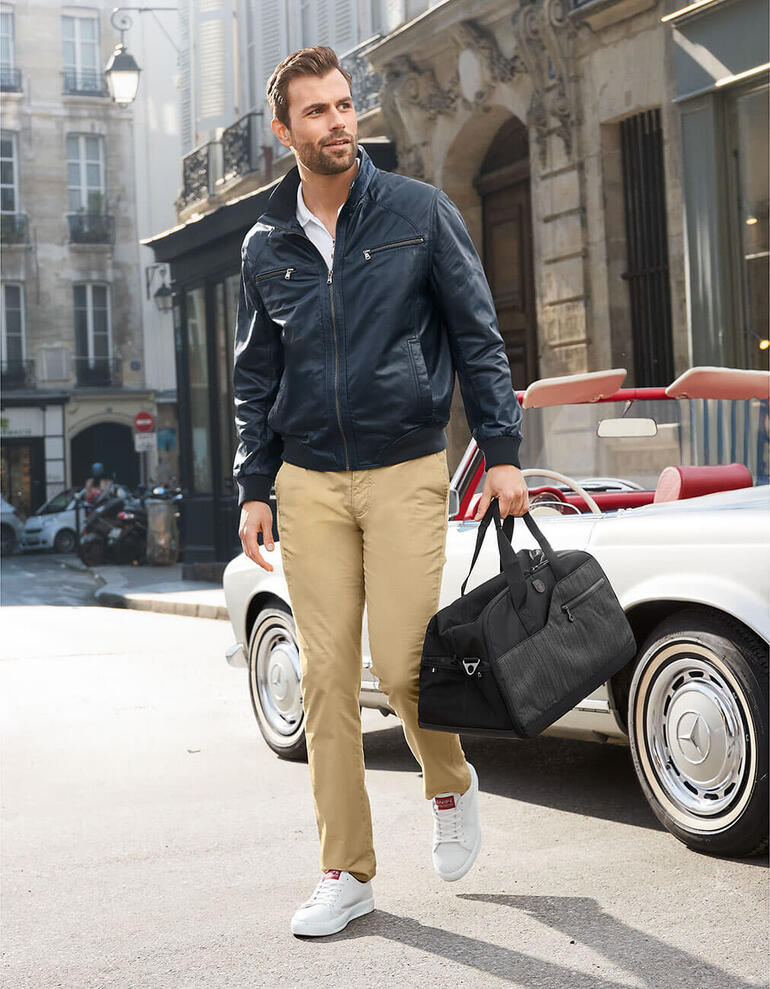 Man in een blauwe leren zomerblouson en beige broek op straat.