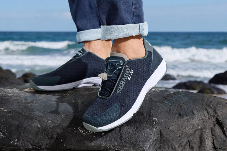 Sebago heren Wet Sneakers in marineblauw, gedragen op een strand met golven op de achtergrond.