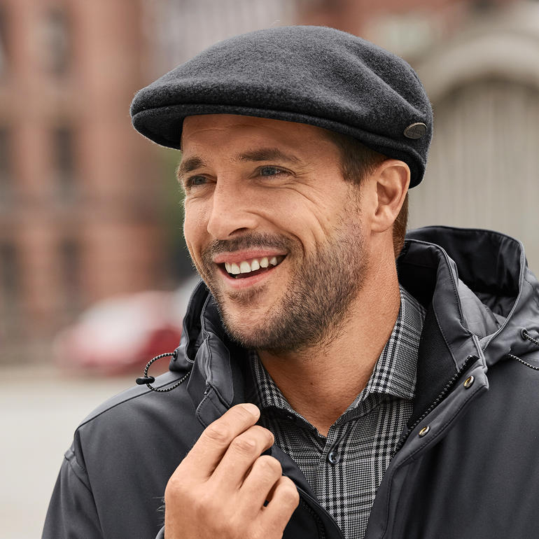 Kangol® flapcap met oorwarmers Man met Kangol-pet en oorkleppen, glimlachend in een stedelijke omgeving.