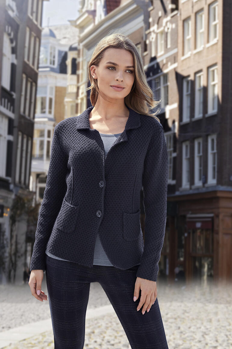 Vrouw in blazer staat voor een schilderachtige stadsachtergrond.