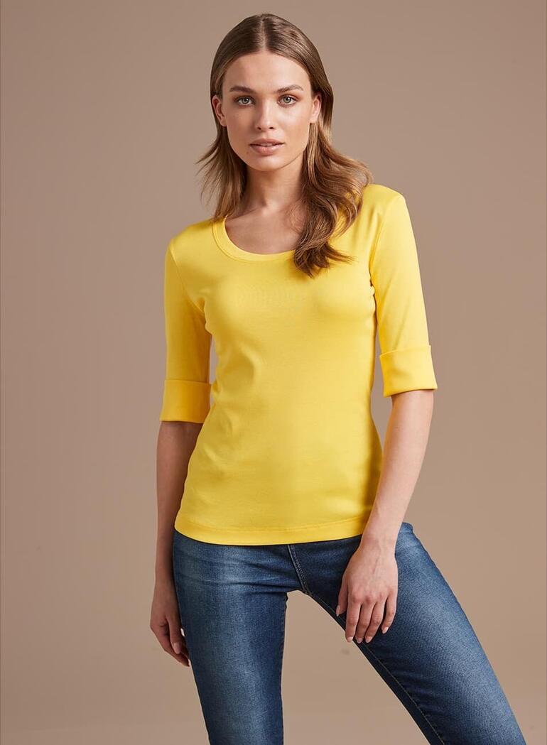 Vrouw met een geel langemouwen shirt en jeans, voor een neutrale achtergrond.