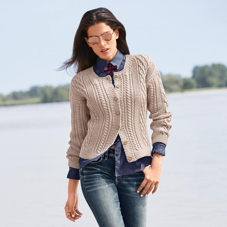 Peregrine Aran-cardigan voor dames Beige cardigan voor dames met kabelpatroon en knopen, gedragen door een vrouw op het strand.